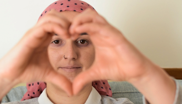 Chica de pañuelo rosado con leucemia haciendo la forma de un corazón con la mano.